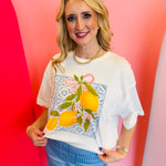 White cotton graphic tee with lemon print, floral frame, and bow accents.