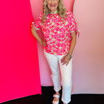Bright pink floral top with green accents, striped neckline trim, and short sleeves with adjustable ties
