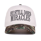 Zenana White cap with black “He Still Does Miracles” embroidery and camo bill, adjustable strap.