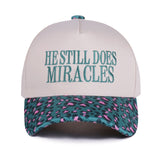 Zenana White cap with turquoise “He Still Does Miracles” embroidery and cougar-print bill, adjustable fit.