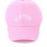 Pink cotton baseball cap with white embroidered “New York” and adjustable brass buckle closure.